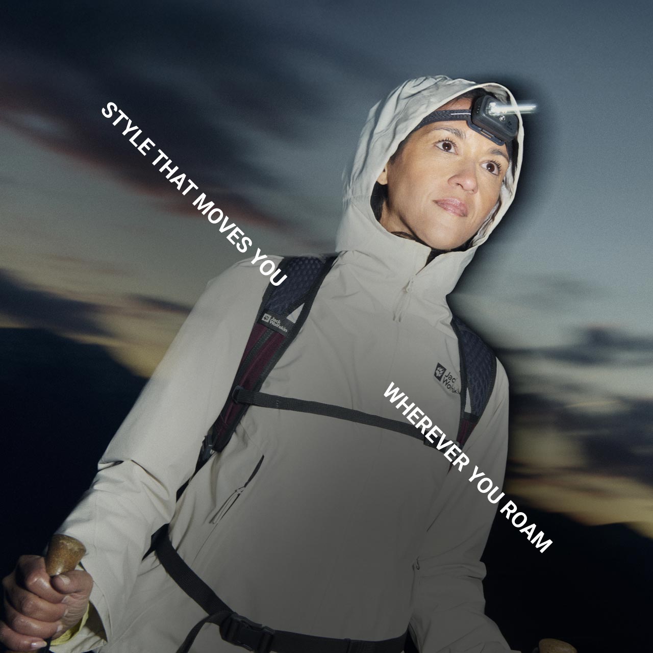 Une personne avec un équipement outdoor et une lampe frontale regarde droit devant et tient des bâtons de trek dans le crépuscule montagneux. Le texte est le suivant : « Style that moves you wherever you roam »