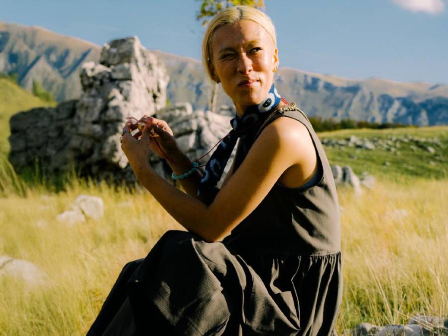Une femme blonde est assise au milieu d'une prairie verte et regarde au loin, entourée de montagnes